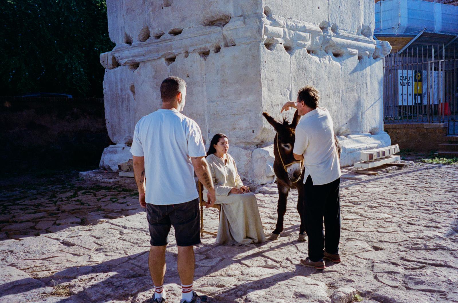 The model seated among the ruins as a handler steadies the donkey in the sunlight